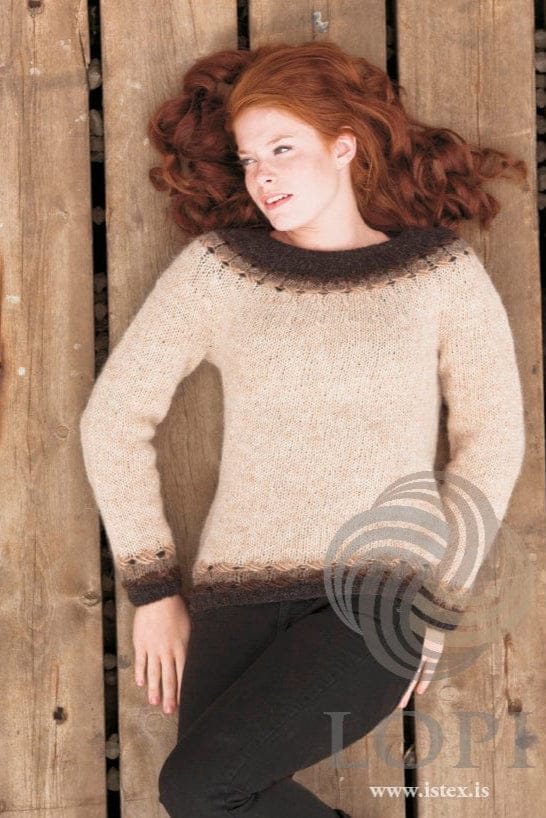 Young woman with long curly red hair lying on weathered wooden boards, wearing Kross in the pattern’s Natural colours — a straw sweater with dark brown rib, brown bands and a row of light crossed stitches above the hem and cuffs, and a yoke with a row of crossed stitches below a wide dark brown top and neck. The black jeans and the small stud earring are not part of the pattern