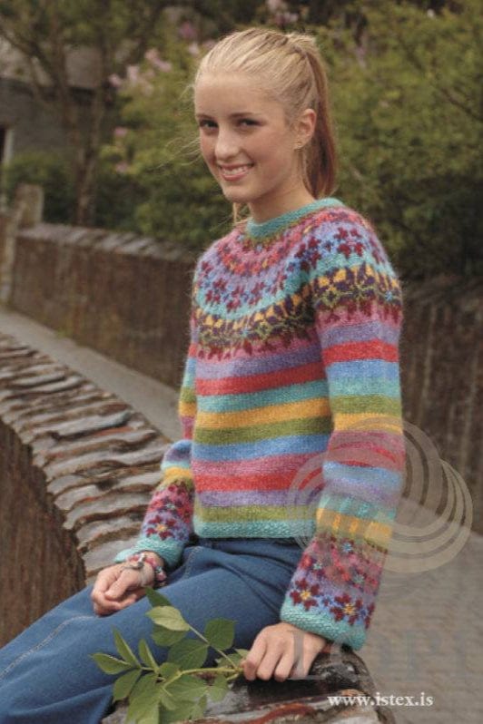 Smiling young woman with a blonde ponytail sitting on a brick wall, bushes behind, wearing Fjör in the pattern’s original colours — stripes of turquoise, green, lilac, coral red, pink, blue and yellow, a yoke and cuff borders of red, violet and pink roses with small flowers, and turquoise moss-stitch edges. The jeans, the bracelets, the stud earring and the leafy twig are not part of the pattern; the LOPI logo and www.istex.is are printed in the corner