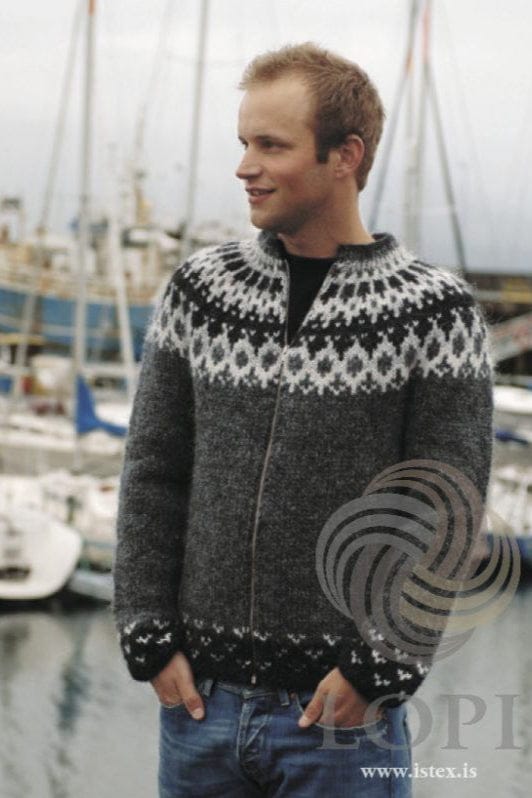 Young man with short reddish-blond hair at a harbour, boats and masts behind, wearing Fugl in the pattern’s own colours — a dark grey cardigan zipped most of the way up, with a yoke of white, ash heather and black diamonds, blocks and bands, and black heather borders with small light V-shapes above the hem and cuffs. The black T-shirt and the blue jeans are not part of the pattern, and the zip is not included. The Lopi logo and www.istex.is are printed on the photo