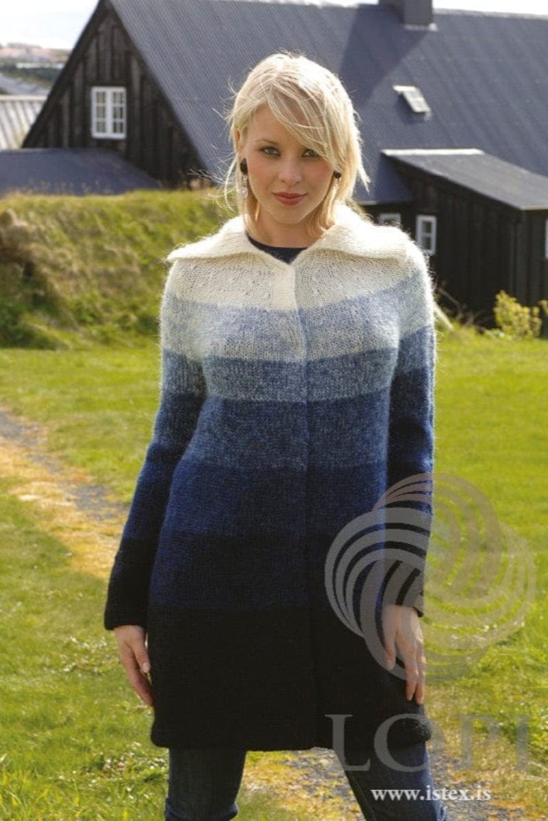 Blonde woman on a lawn in front of black timber houses and a grassy bank, wearing the Blue version of Land in the pattern’s colours — a long jacket shading in bands from black at the hem through dark and lighter blues to white at the shoulders, with a deep white rib collar folded over and the front closed. Denim and faded denim, two of the blues, are no longer made. The earrings, the dark top and the dark jeans are not part of the pattern