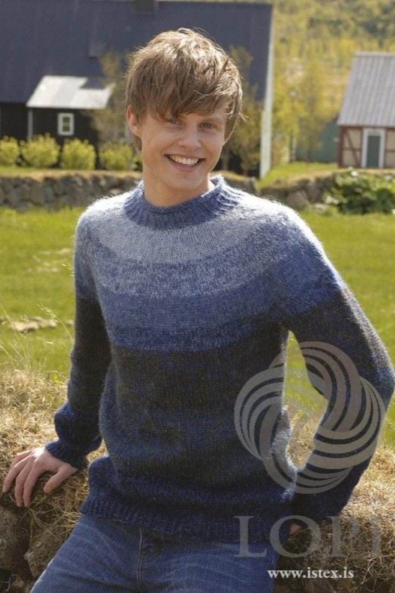 Smiling young man sitting on a grassy bank, houses and a garden behind, wearing the Blue version of Útjörð in the pattern’s colours — a round-yoke sweater, dark blue at the hem, shading up in bands of blue to a pale grey-blue yoke, with a blue ribbed neckband. Sea green, denim and faded denim, three of its colours, are no longer made. The blue jeans are not part of the pattern
