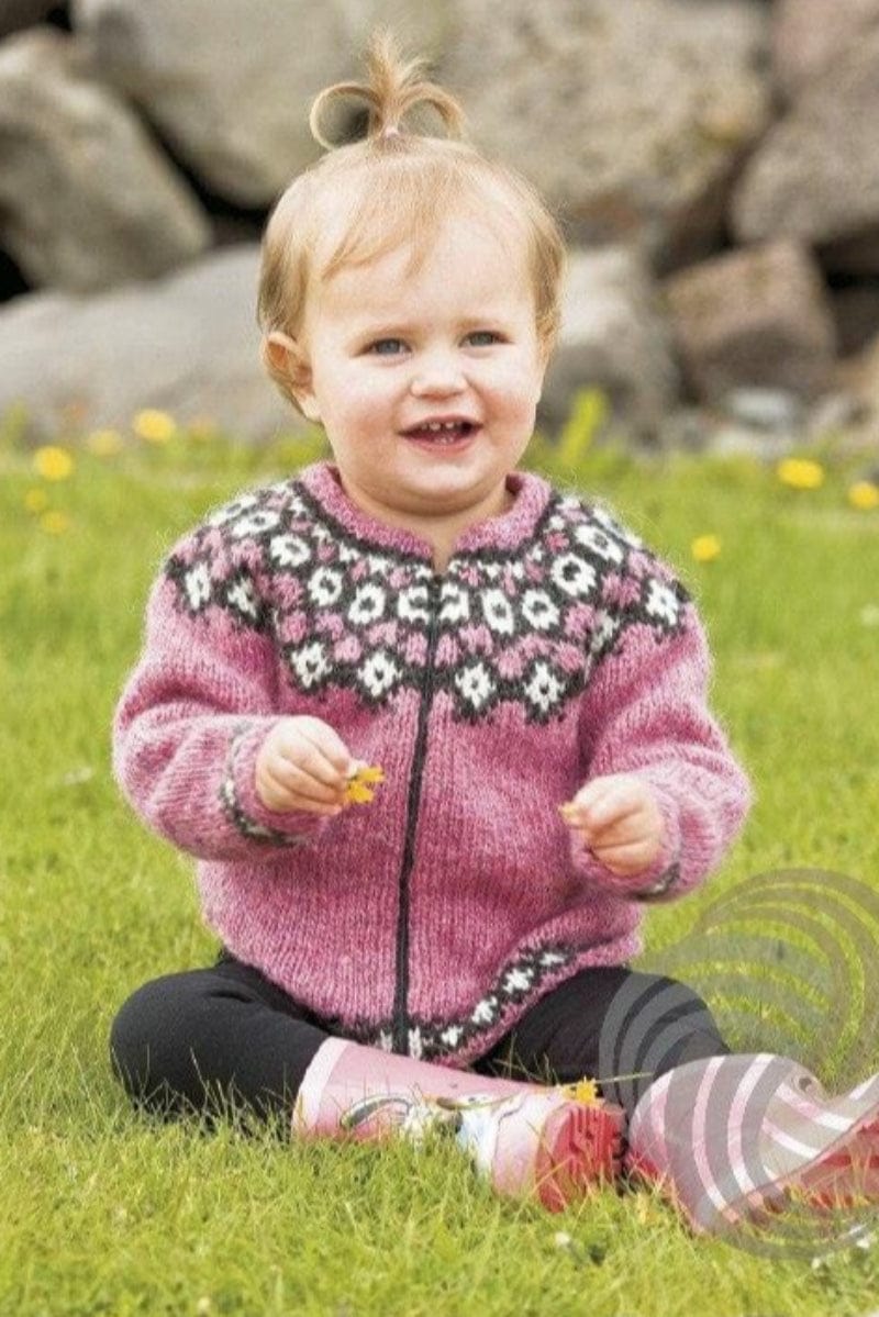 Smiling toddler with a little topknot sitting on grass with yellow flowers, rocks behind, wearing the pink Kría cardigan in the pattern’s own colours — pink, zipped with a dark zip, with a yoke of white motifs on black heather and pink, and black heather and white bands above the hem and cuffs. The small pink hair tie, the black leggings and the pink rubber boots are not part of the pattern, and the zip is not included