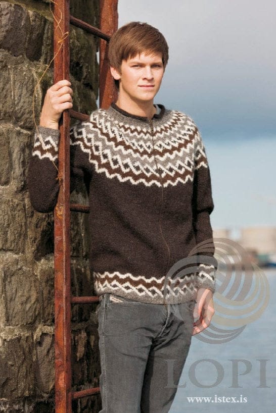 Young man with brown hair leaning on a rusty iron ladder against a dark stone wall, water behind, wearing Hlekkur in the pattern’s Black version — a black sheep (dark brown-black) zip cardigan with a yoke of chocolate, white and grey zigzag bands and small dark diamonds, the same zigzag border above the grey rib at the hem and cuffs, and a dark roll at the top of the neckband. The grey jeans and the light top showing at the neck are not part of the pattern