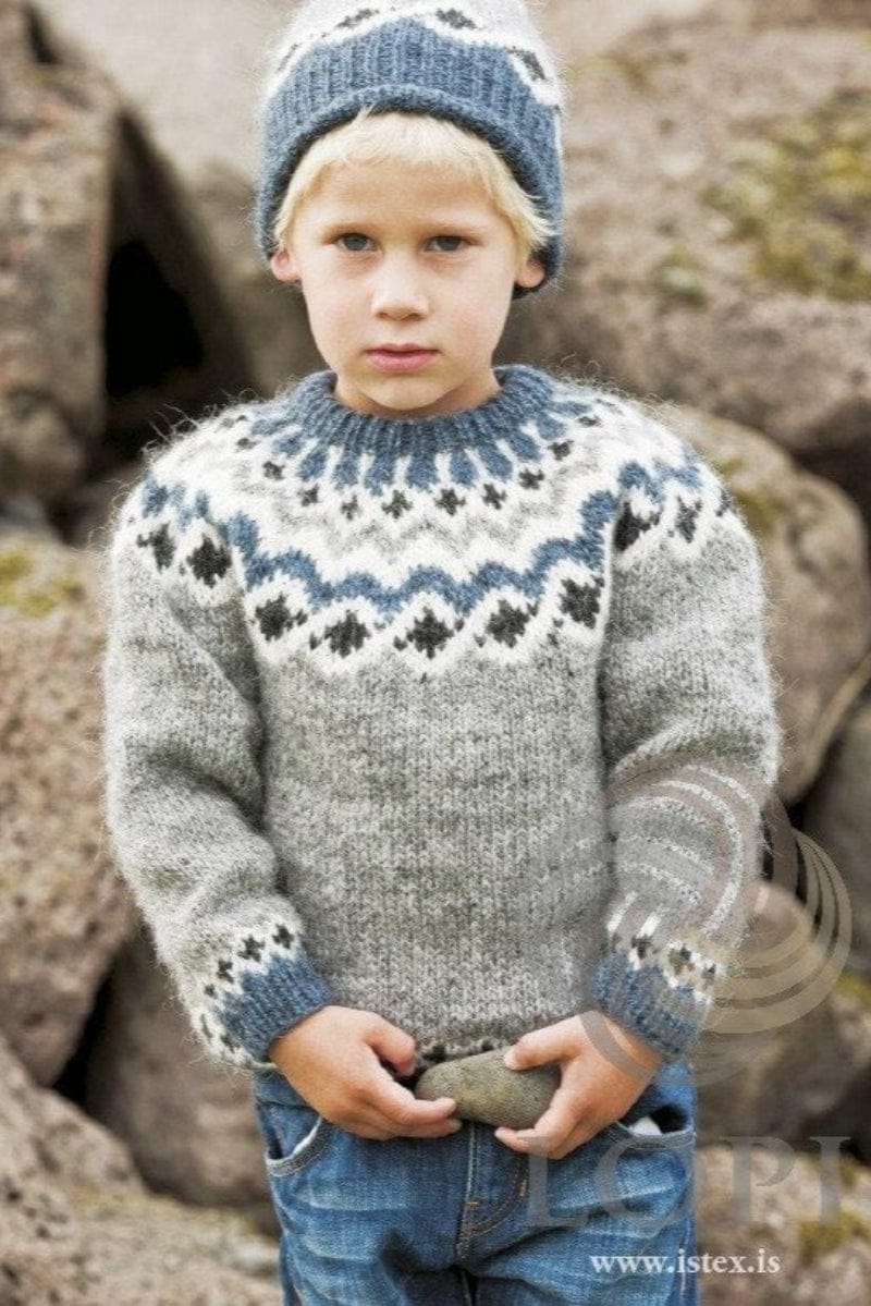 Fair-haired boy among large rocks in the Grey version of Kambur, in the pattern’s own colours — a light grey sweater with a white yoke of black heather diamonds, a stone blue wave, grey zigzags and small black heather crosses, stone blue at the neck, and a border of small crosses above the stone blue cuffs — and the matching hat, its stone blue rib turned up, with black heather diamonds on white. The blue jeans and the stone in his hands are not part of the pattern