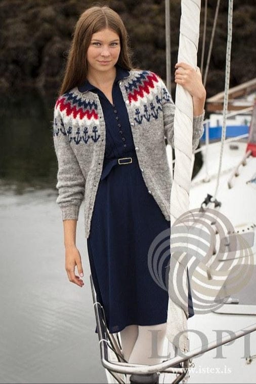 Young woman on the bow of a white sailing boat in the Ankeri cardigan, shown in the pattern’s own colours — light grey Einband with a band of navy anchors at the base of the yoke and zigzags of white, flame red, cardinal, grey and navy above it, worn open, with white buttons. This photo is from Ístex’s photo set for the pattern, not the PDF. The navy dress with its belt and the white tights are not part of the pattern, and the buttons are not included