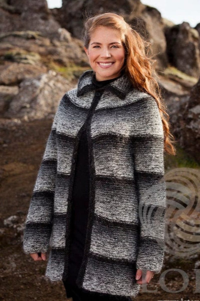 Smiling woman with long red hair among tall mossy rocks in the greytone Katla, shown in the pattern’s own colours — a long garter-stitch cardigan in bands of white and greys with darker bands between, the collar folded down and black heather button bands. The black polo neck and the black clothes under the cardigan are not part of the pattern