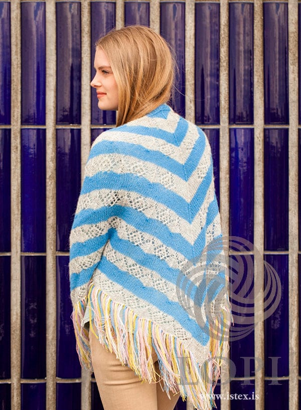Back view of Vængur over a young woman’s shoulders against dark blue tiles, in the pattern’s own colours — the sky blue and ash-heather lace stripes meeting in a point at the centre back, with the fringe below. The beige trousers are not part of the pattern
