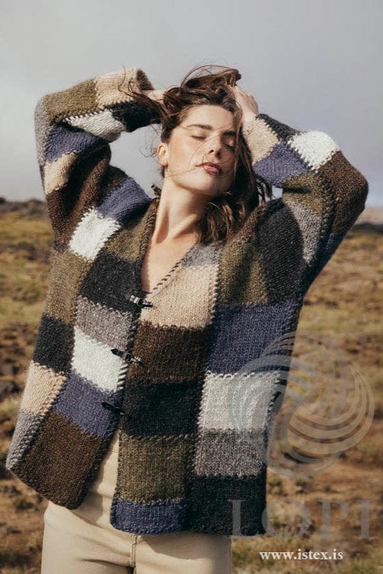 Woman with her hands in her hair on brown moorland wearing the earth-tones Brautir, shown in the pattern’s own colours — a long V-neck cardigan of staggered blocks in black heather, indigo, highland green, ash, grey, wheat and chocolate, with seed-stitch ridges between them and three dark hook-and-eye clasps on the outside. The beige trousers are not part of the pattern