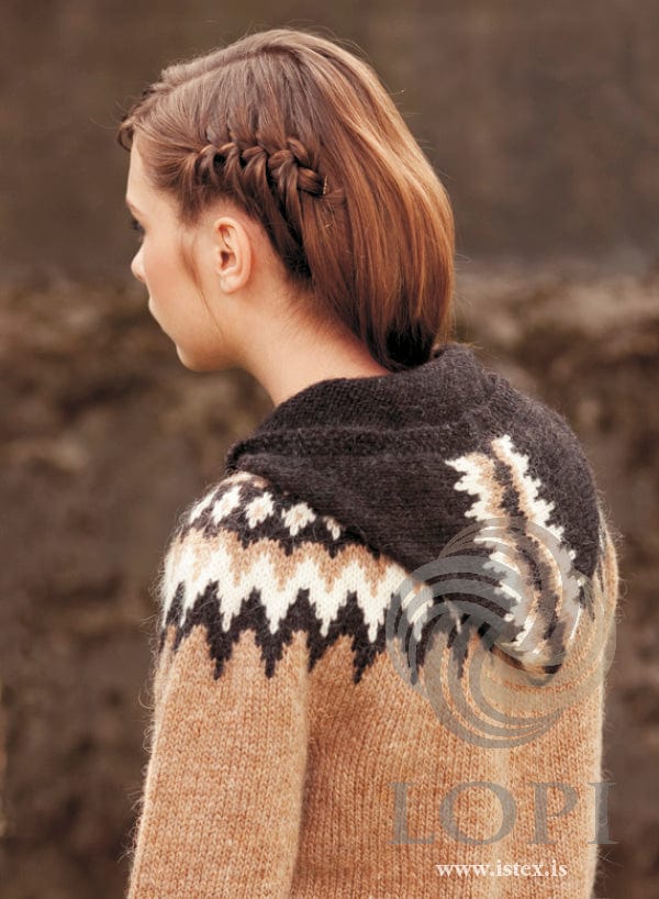 The same young woman seen from behind and to the side, her hair in a braid, showing the brown Var in the pattern’s own colours — the black-sheep hood lying on her back with its band of white and barley shapes, and the barley yoke with black-sheep and white zigzags. The hair grip in her braid is not part of the pattern