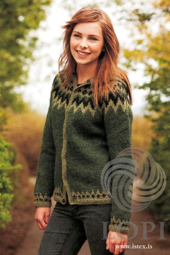 The same young woman smiling on a path between trees in the green Var, shown in the pattern’s own colours — a pine green cardigan with a yoke of black zigzags on celery green, celery green moss-stitch bands and border, and a press stud showing at the hem. The dark top at the neck and the grey jeans are not part of the pattern