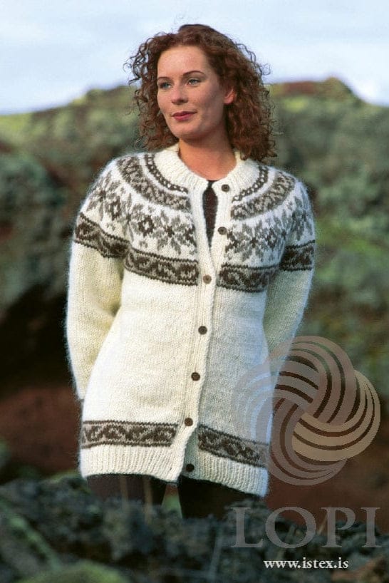 Woman with curly red hair outdoors, hands behind her back, in the Lopi 18 no. 8 cardigan, buttoned, shown in the pattern’s own colours — white with a yoke of oatmeal stars and brown acorn diamonds between two acorn and light beige bands, a matching wave border above the ribbed hem and cuffs, ribbed front bands and small round brown buttons. The dark top under it and her dark tights or trousers are not part of the pattern; the buttons are not included