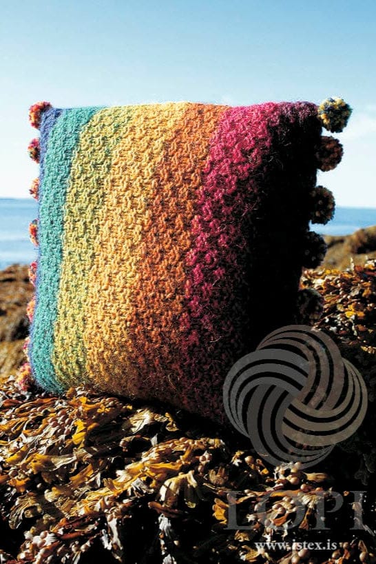 Þúfa standing on seaweed-covered rocks, the sea and a blue sky behind — a square cushion in the pattern’s colours, striped from sapphire blue at one edge through teal, chartreuse green, golden, amber and fuchsia to bordeaux at the other, in textured double moss stitch, with small multicoloured pompoms along the two sewn edges. Sapphire blue heather is no longer made by Ístex