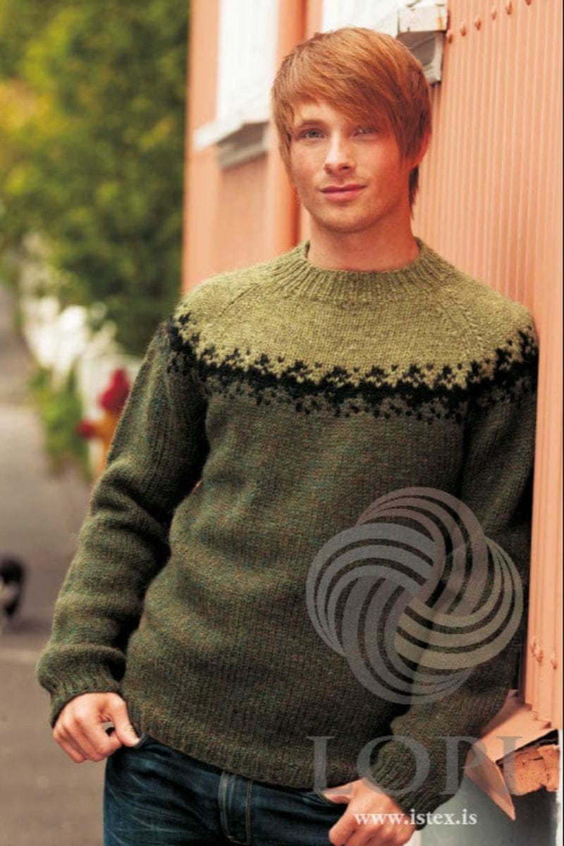 Young man leaning on a salmon-pink wall in the green Sátt, shown in the pattern’s own colours — a dark spruce green Plötulopi sweater with a black jagged band at the base of the yoke and a light clover green upper yoke with raglan lines and a ribbed neckband. The blue jeans are not part of the pattern
