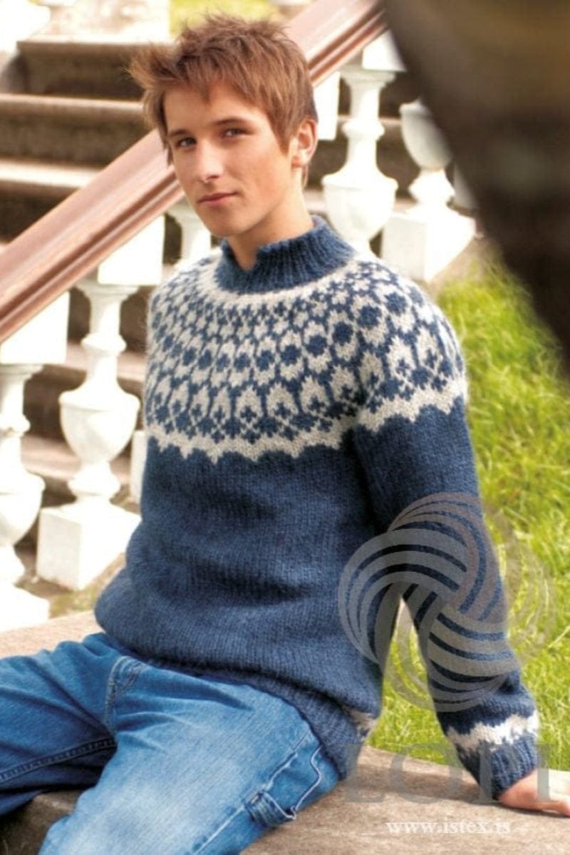 The same young man sitting on a stone ledge in front of steps with white balusters and a wooden handrail, wearing the blue Virki in the pattern’s own colours — indigo blue with an ash heather yoke and cuff border and an indigo blue ribbed collar. The jeans are not part of the pattern; the LOPI logo and www.istex.is are printed in the corner