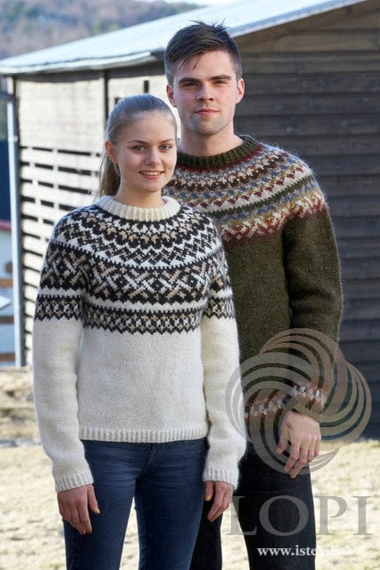 Young woman and young man in front of a dark wooden shed in two Sjóna versions, shown in the pattern’s original colours — she in the white sweater with a black sheep and straw yoke and a matching band just below it; he in the moss green sweater with a yoke of narrow bands in rust, stone blue, golden, beige, brick and browns and a band above the cuffs (its hazel brown is no longer made). Her blue jeans and his dark trousers are not part of the pattern