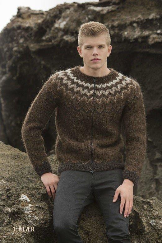 Young man sitting on a rock in front of a dark lava cliff in the brown Blær, shown in the pattern’s own colours — a chocolate Álafosslopi zip cardigan with a yoke of zigzag bands running from dark at the bottom to white near the neck, and the men’s dark zigzag border and dark rib at the hem and cuffs. The dark grey trousers are not part of the pattern