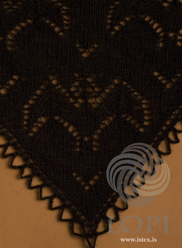 Close-up of Blaka against a brown background, in the pattern’s own black Einband — the eyelet outlines of the lace near the point, and the crocheted chain loops along both sloping edges where they meet at the point