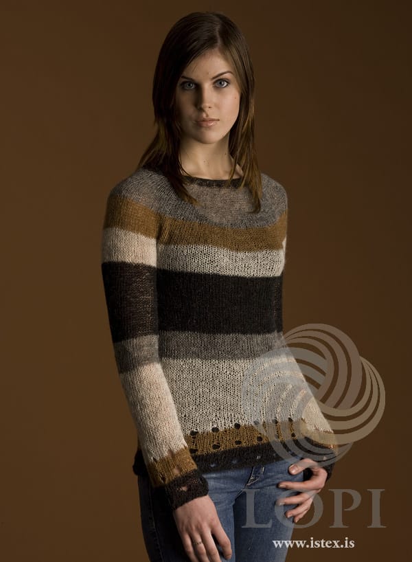 Woman in Hraði, shown in the pattern’s own colours — a light Einband round-yoke sweater in wide stripes: black with a round of holes at the hem, tawny with holes, a long ash stripe, dark grey, a wide black stripe and ash at the chest, then a tawny band and grey in the yoke up to a narrow black neck edge; the sleeves are striped the same way. The jeans and her ring are not part of the pattern