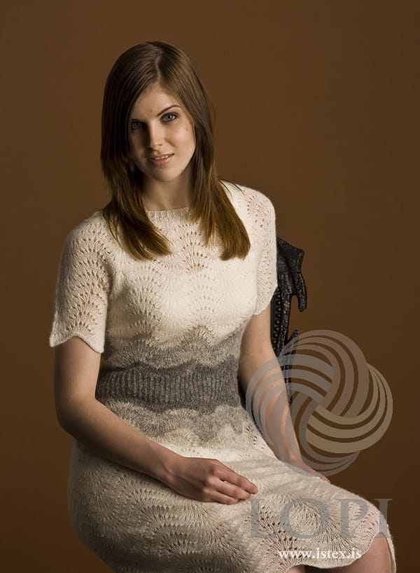 Woman seated in Miðja, the white and grey version with short sleeves, in the pattern’s own colours — the round lace yoke, grey waves shading into the twisted-rib waist, and the lace skirt over her knees. The dark carved chair is not part of the pattern