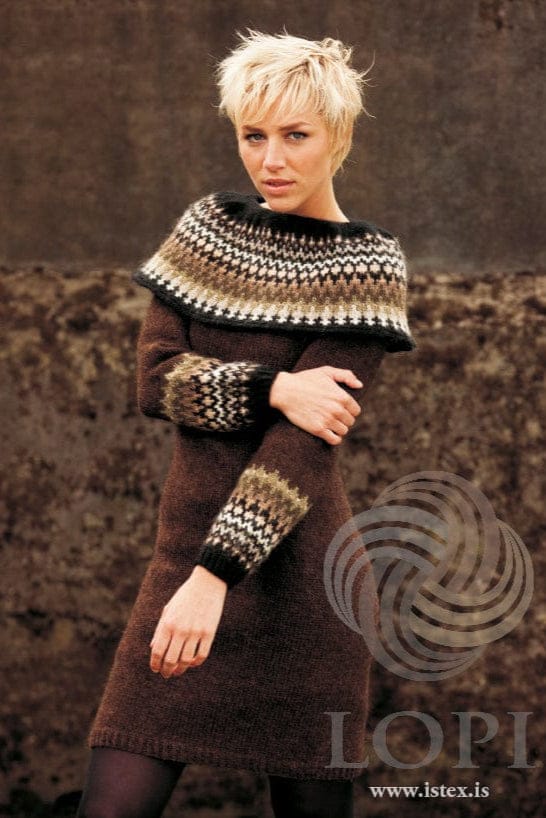 Young woman against a dark brown background in the brown Hvarf dress, shown in the pattern’s own colours — a chocolate Léttlopi dress with a black-edged cape collar of small motifs in white, light beige, straw, olive and brown shades, and black cuffs below a matching band. The dark tights are not part of the pattern