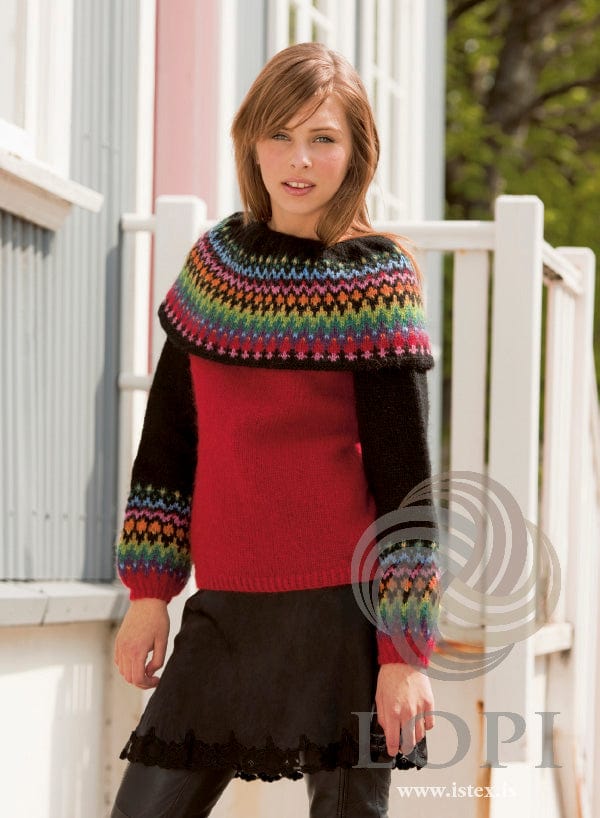 Young woman in front of a pale house and a white railing in the red Hvarf sweater, shown in the pattern’s original colours (orange and mustard are no longer made) — a crimson Léttlopi sweater with black sleeves, crimson cuffs below a band of bright colourwork, and a wide black-edged cape collar with rows of small motifs in pink, red, orange, yellow, green, teal, violet and blue. The black skirt with a lace hem and the black leather-look trousers are not part of the pattern