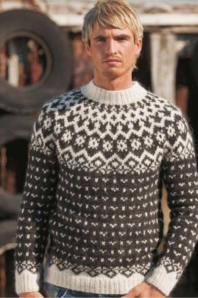 Fair-haired man in front of a stack of old tyres, wearing Vetur in the pattern’s own black and white — black heather body and sleeves covered in small white motifs, a white border of points above the white rib, and a yoke of small diamonds and white rosettes rising to white with small black motifs at the ribbed neck. The light blue jeans are not part of the pattern