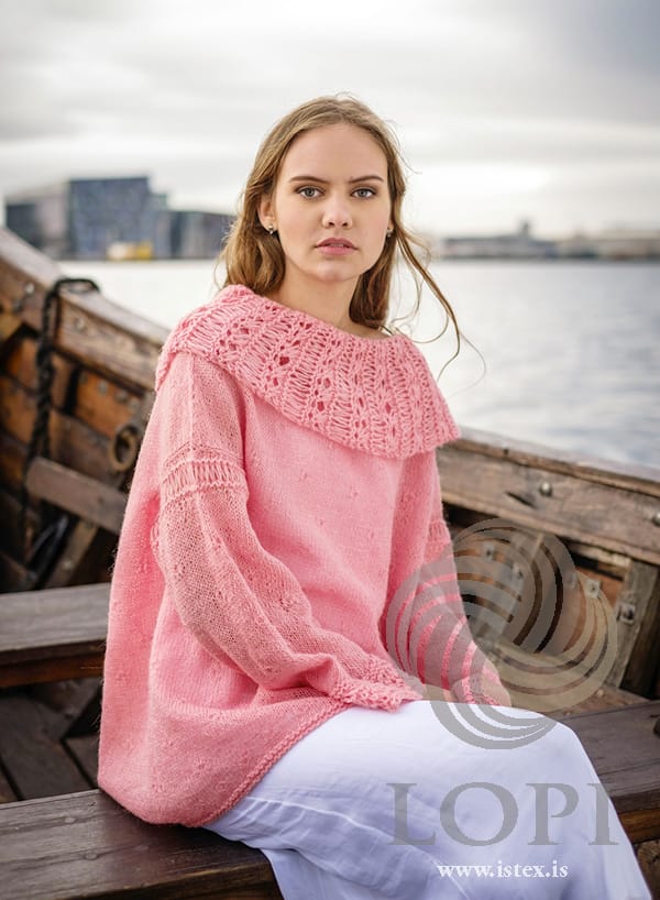 Woman sitting on the side of a wooden boat in Askja — a loose blush Einband sweater with a deep openwork collar, small diamonds scattered over the body and sleeves, a band of long stitches on each upper arm and wide openwork cuffs, in the pattern’s original colour. The white dress and earrings are not part of the pattern