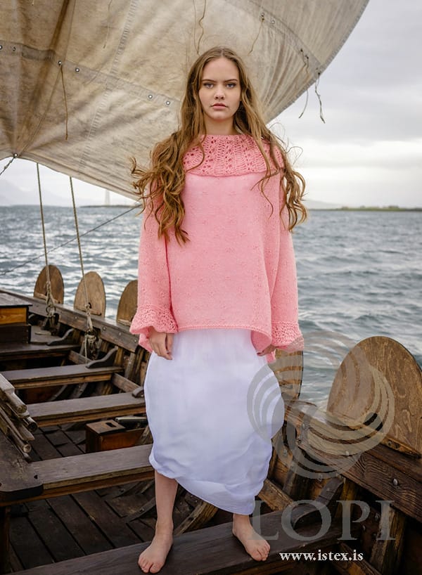 Askja from the front, worn standing on a wooden boat under a sail — loose, with the deep collar and wide openwork cuffs, in the pattern’s original blush. The long white dress is not part of the pattern