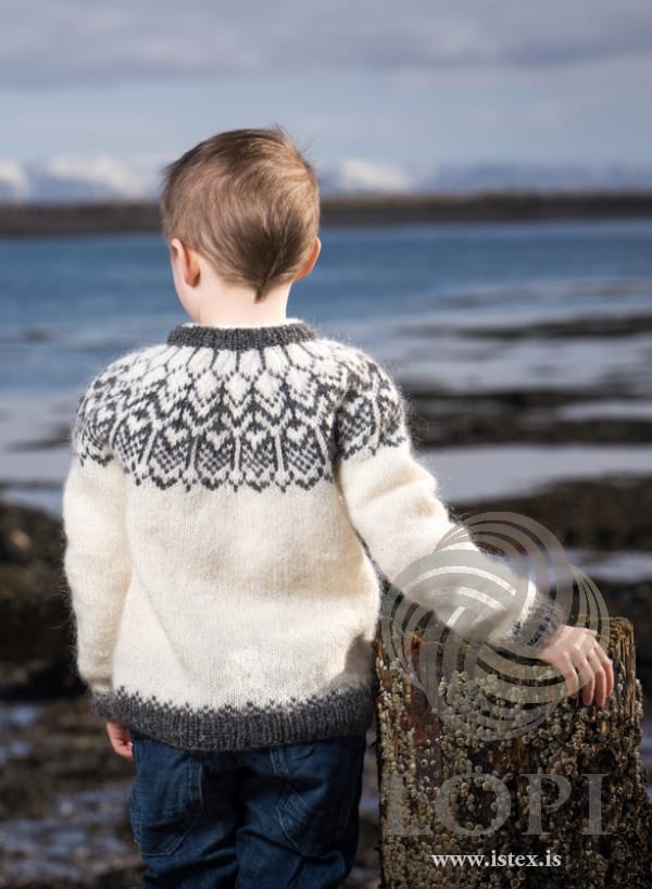Fura from the back on a boy at the shore, one hand on a wooden post — the grey yoke pattern, the grey neckband and the check band above the hem rib. The jeans are not part of the pattern