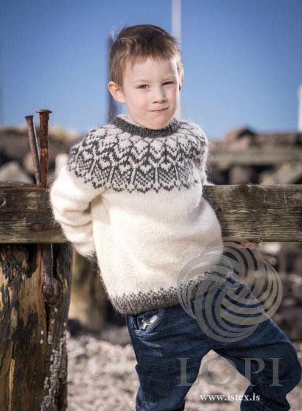 Boy leaning on a weathered wooden fence in the white and grey Fura yoke sweater. The jeans are not part of the pattern