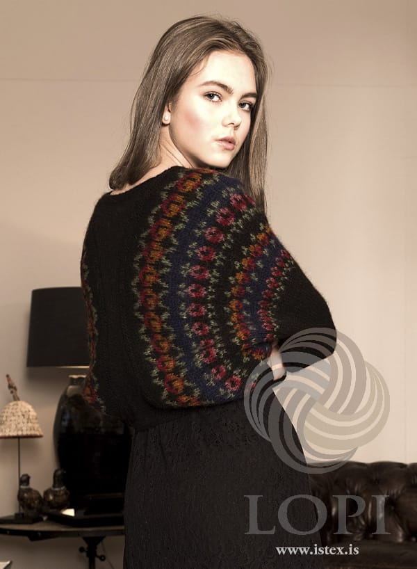 Heartwarmer in the red roses colourway, from the back — roses in garnet red, rust and pink with sage green and a navy band, arching over the back and arm, shown in the pattern’s original colours (sage green and rose heather are no longer made). The long black lace dress is not part of the pattern