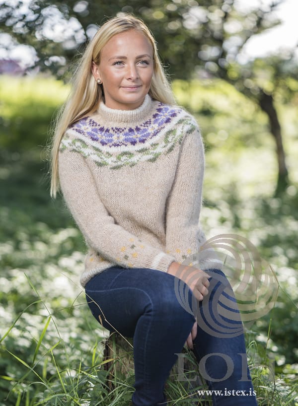 Woman sitting outdoors in Manitoba — a straw Léttlopi pullover with a round yoke of violet and lilac flowers above a band of green leaves, small yellow and green dots above the white-striped rib at the hem and cuffs, and a folded neckband, shown in the pattern’s original colours (sun yellow and lilac are no longer made). Her jeans and earrings are not part of the pattern
