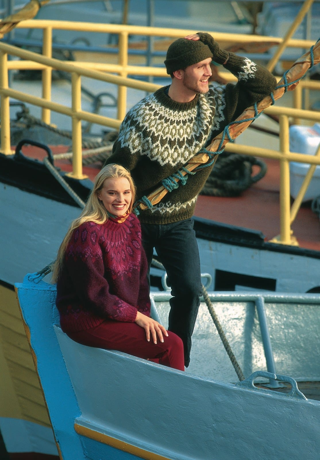 Smiling woman on the bow of a boat in the Lopi 12 no. 7 sweater — a dark plum body, raspberry rib at the hem and cuffs, and a raspberry yoke with pink ovals ringed in plum and raspberry rays up to the neck, shown in the pattern’s original colours in an old film photo (all three are no longer made). Her scarf and red trousers are not part of the pattern; the man behind her wears a different pattern