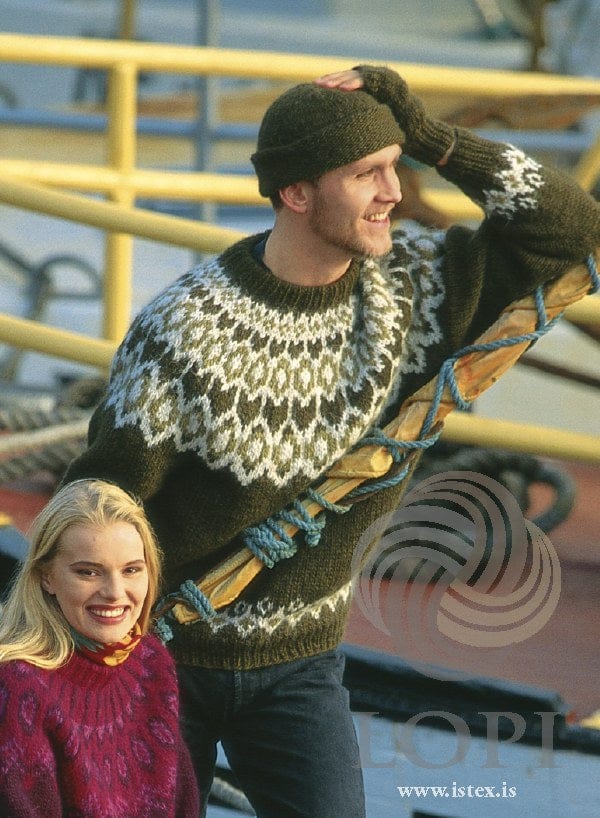 Smiling man on a ship’s deck in the Lopi 12 no. 9 sweater, the rolled-edge hat and a fingerless glove — dark olive green with a deep yoke of pale grey scallops and honeycomb shapes filled with khaki and dark green, and the same border above the hem (shown in the original colours — this kit has cypress green heather for the loden green 9239, and highland green, darker and browner, for the golden green 9240). The woman’s pink sweater and scarf, and the man’s jeans, are not part of the pattern
