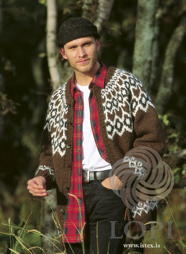 Man at the edge of a birch wood in the Lopi 12 no. 10 cardigan, worn open — chocolate brown with a deep yoke of white and black heather motifs, a matching border on the cuffs and above the hem, and buttons down ribbed bands — and the rolled-edge hat from the same leaflet; the plaid shirt is not part of the pattern (shown in the original colours — this kit has black tweed for the hat’s 9239 tweed)