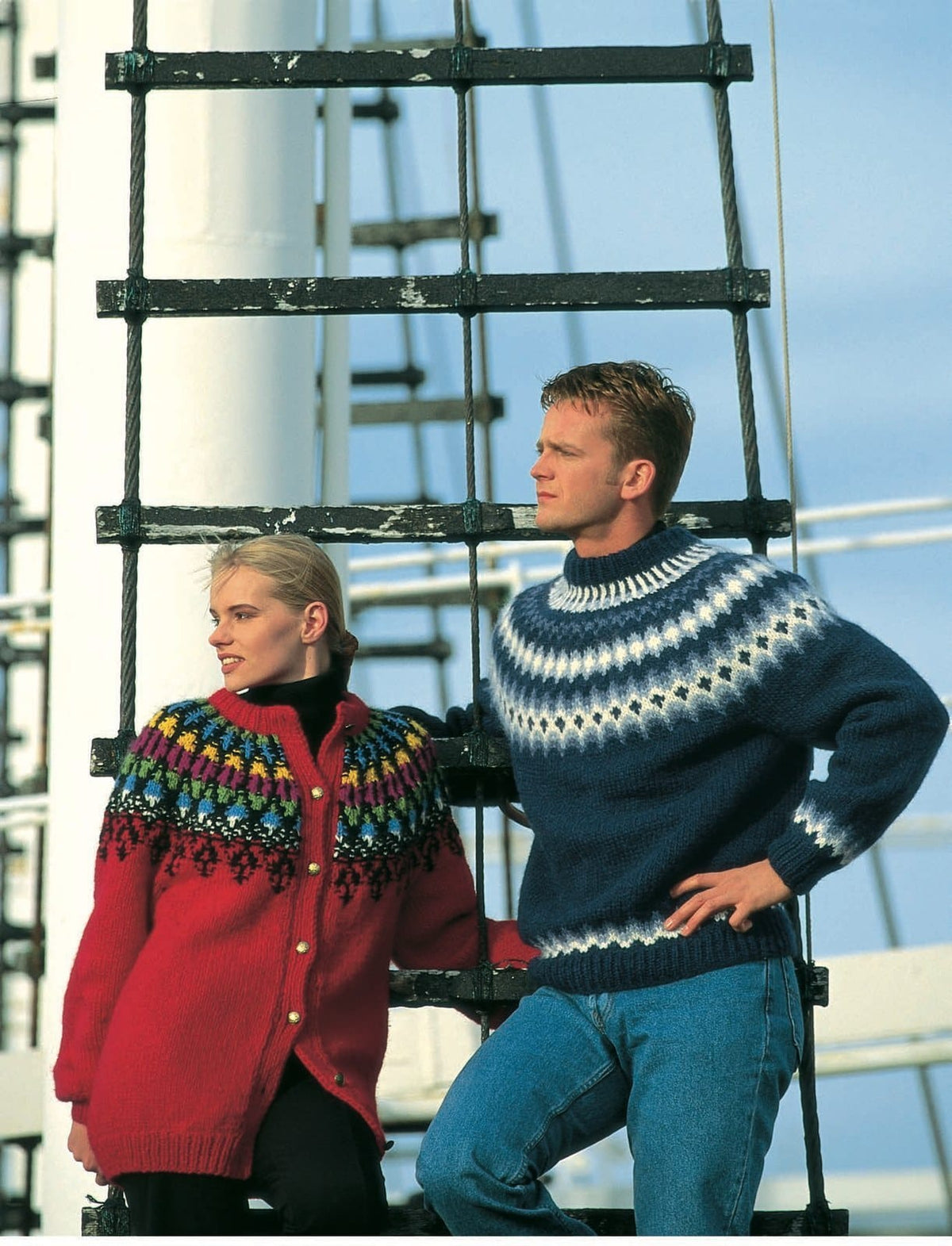 Woman by a ship’s rope ladder in the Lopi 12 no. 17 cardigan — long and scarlet red with gold buttons, and a textured yoke: black motifs rising into the red, a black and white band, blue tulip shapes, then green, fuchsia, yellow, blue and green rounds up to a red neckband, shown in the pattern’s original colours (all but the black and white are no longer made). Her black polo neck and trousers are not part of the pattern, nor is the man — he wears Lopi 12 no. 19, a different pattern, and jeans
