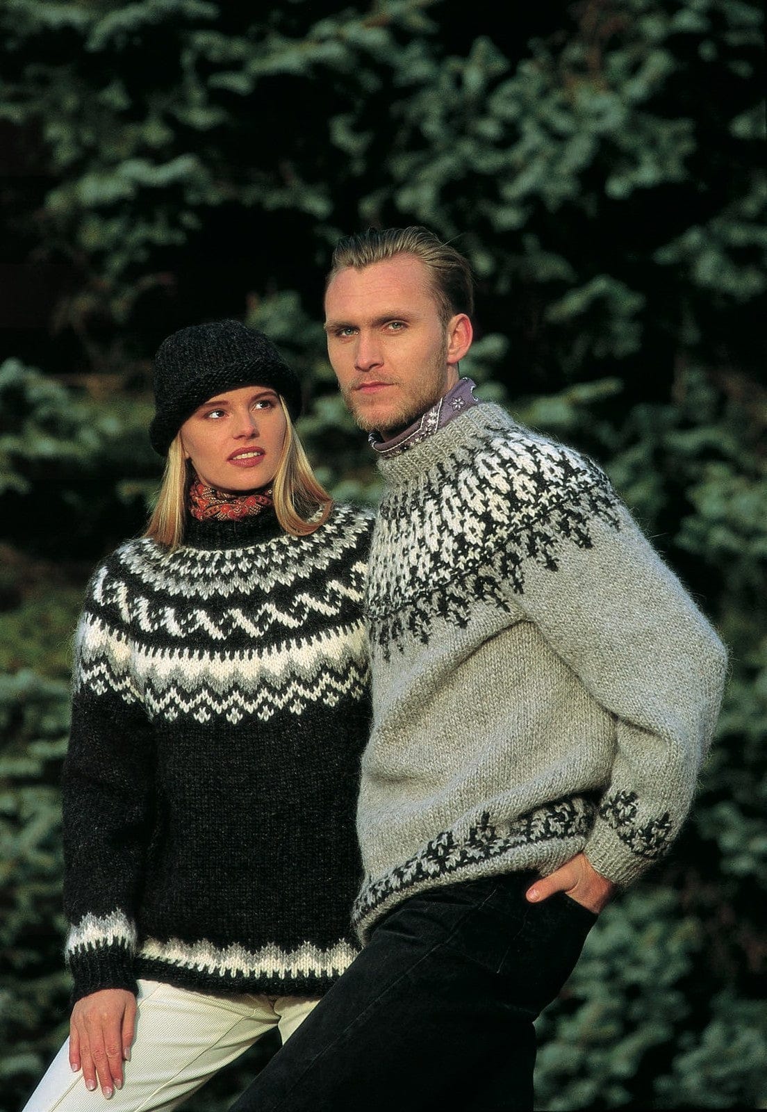 Man in the Lopi 12 no. 18 sweater in front of fir trees — a light grey ash heather body, black heather motifs on the yoke rising into a white band near the neck, and a black border above the hem and cuffs, shown in the pattern’s own colours. His lilac neckerchief and black trousers are not part of the pattern. The woman wears Lopi 12 no. 14, a different pattern; her black hat, red scarf and light trousers are not part of this one either
