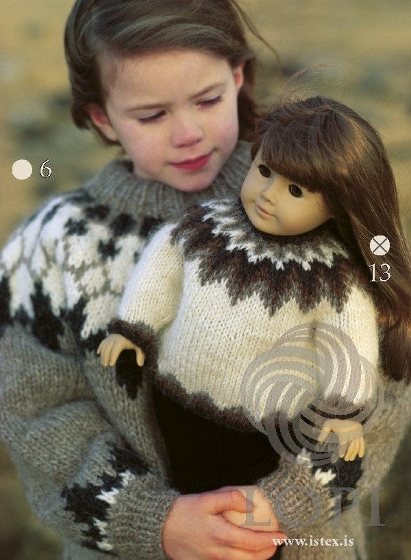 A girl holds a doll in Design 13 from the same leaflet — the white doll sweater with brown and near-black zigzags in the yoke and dark ribs, sold as a separate kit (Lopi 18 No. 13). The girl’s grey sweater is another pattern (Lopi 18 no. 6)