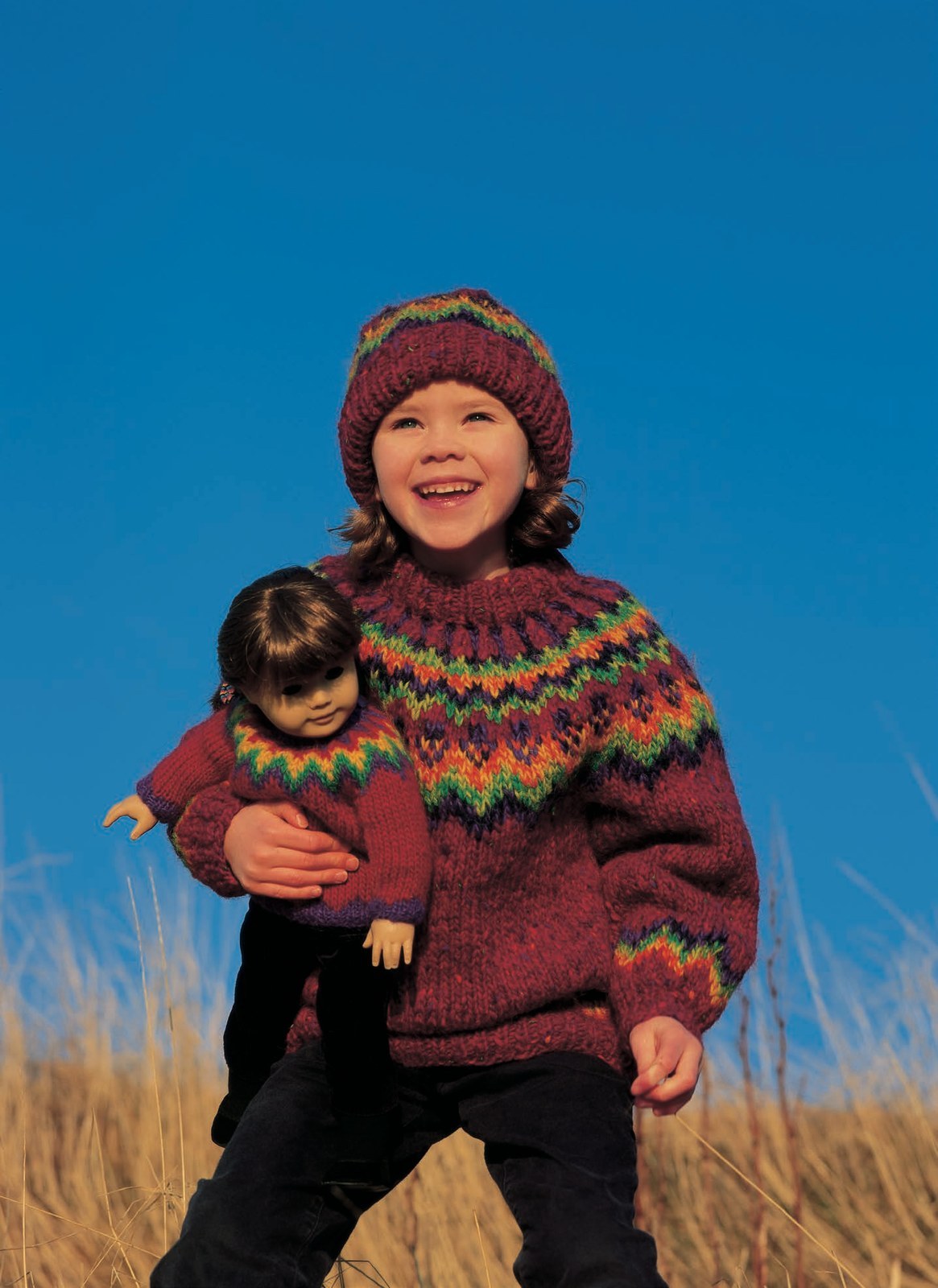 Design 12 from the same leaflet — the crimson red doll sweater with a yoke of purple, green and yellow zigzags, carried by a laughing child; sold as a separate kit (Lopi 18 No. 12). The child’s own sweater and hat are another pattern