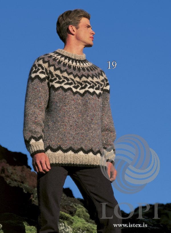 Man on a mossy hillside in the Lopi 18 no. 19 sweater — grey with a yoke of a black zigzag, black slanting leaves on cream, black triangles and black spikes up to the folded light beige neckband, and a black zigzag above the light beige hem and cuffs (shown in the original colours — this kit has plain grey heather, without the coloured flecks, for the gray fleck, and light ash heather for the ash); the dark trousers are not part of the pattern