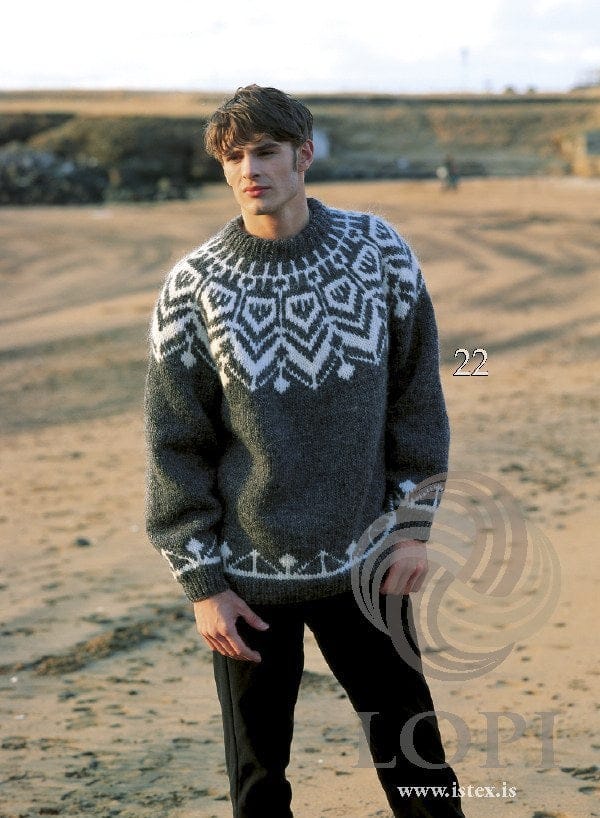 Man on a beach in Design 22 of this pattern, in its own colours — dark gray with a white yoke of large pointed shapes, zigzags and small diamonds, a white border of diagonal lines and small trees above the hem and cuffs, and a folded rib neckband. His black trousers are not part of the pattern
