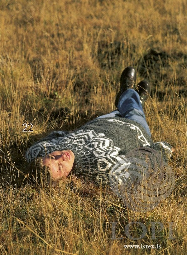The same dark grey sweater seen from above, worn by someone lying in long grass, showing the white yoke; the jeans and boots are not part of the pattern