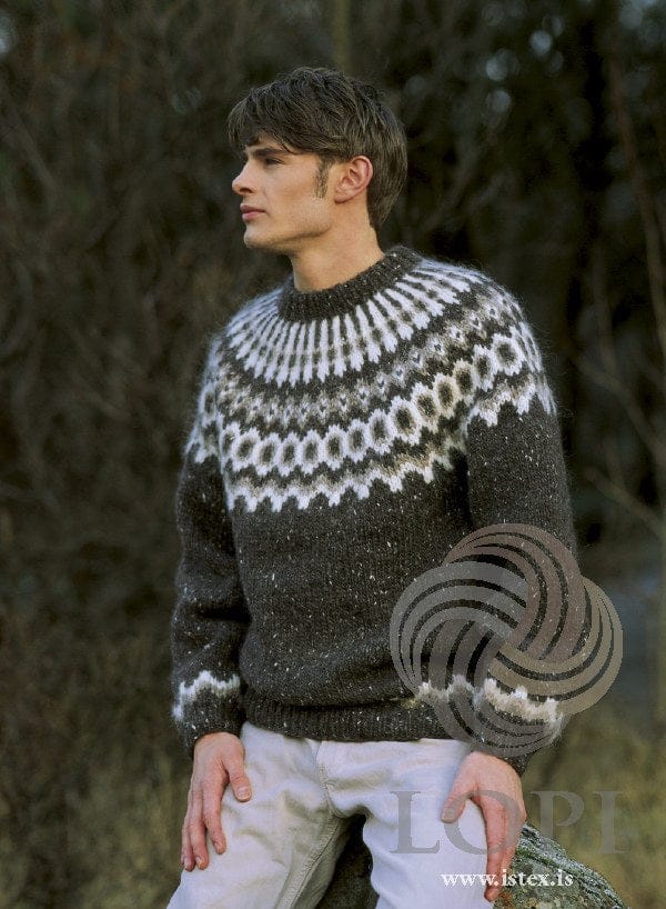 Man seated on a mossy rock in the Lopi 18 no. 28 sweater, shown in the pattern’s original colours — black sheep tweed, a very dark brown-black with small white flecks (9452, no longer made), with a white and oatmeal yoke of a zigzag band, a row of rings, small motifs and white stripes rising to the neck, and a zigzag border above the cuffs. His pale trousers are not part of the pattern