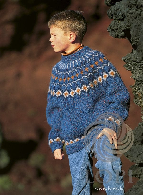 Boy by a lava rock in the Lopi 18 no. 30 children’s sweater — blue tweed with a yoke of pale diamonds with orange centres outlined in navy, small orange crosses and pale spikes up to the folded neckband, and a matching border above the hem and cuffs (shown in the original colours — this kit has blue tweed with white flecks for the monet with its coloured flecks, and amber heather, darker and rustier, for the copper); the orange collar of the top under it and the jeans are not part of the pattern
