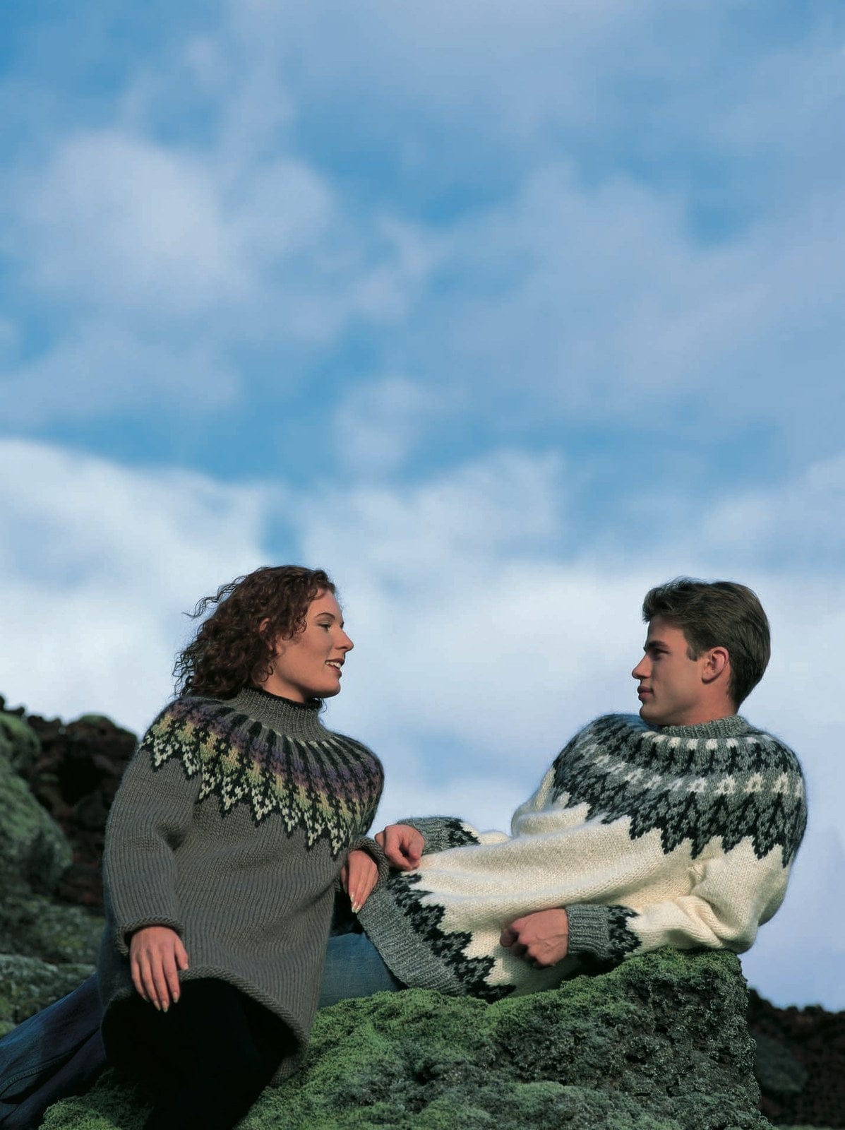 A man and a woman on a mossy lava rock under a blue sky. On the right, the man wears the Lopi 18 no. 41 sweater — white, with a light grey round yoke of black spikes and zigzags, a band of small white diamonds and black and white stitches up to the folded grey neckband, and a black zigzag border above the grey ribs at the hem and cuffs. On the left, the woman wears Design 40 from the same leaflet, which is not available as a kit; the jeans and trousers are not part of the pattern