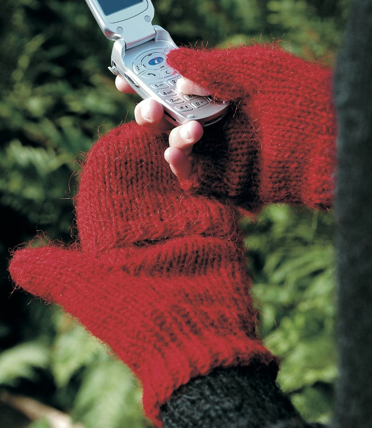 Two hands in the crimson red GSM mittens — one with the mitten top folded back, the fingertips out and the thumb tip through the thumb opening on the keys of a phone; the other with the top pulled over the fingers