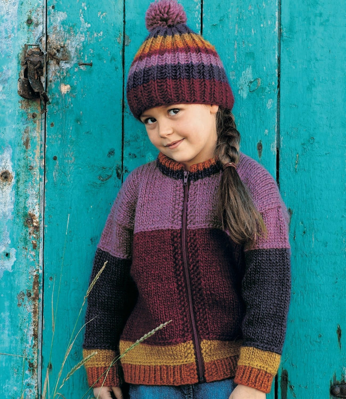 The same Haddur hat in its burgundy version — burgundy, purple, orchid pink, rust and ochre stripes with an orchid pompom — worn with the burgundy Hlökk jacket. The burgundy version is not sold as a kit: four of its five colours are no longer made