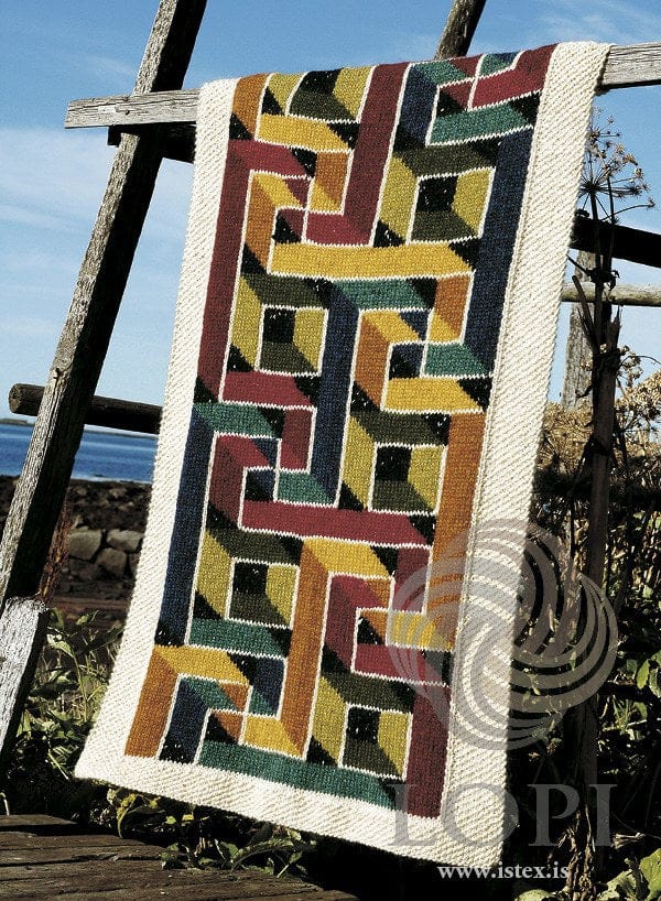 Ravenna hung over an old wooden frame outdoors, with the sea and sky behind and plants at the side, in the pattern’s original colours (sapphire blue heather is no longer made) — bands of golden, amber, ruby red, fuchsia, deep blue, teal, chartreuse, cypress green and black tweed crossing over and under each other, each outlined in ecru, inside a wide ecru moss-stitch border. The wooden frame is not part of the pattern
