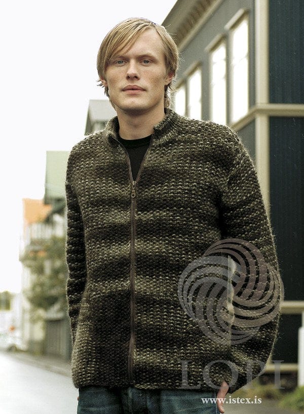 Man in a street of old houses wearing Rökkur from Lopi 24, shown in the pattern’s original jungle green (9980, no longer made), which knits up in uneven olive and near-black bands — moss stitch all over, no rib at the hem or cuffs, a high stand-up collar and a front zip, here open to mid-chest. The black T-shirt and the jeans are not part of the pattern