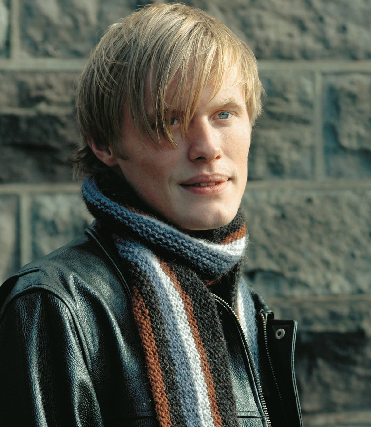 Young man in a black leather jacket wearing Vegur — a garter-stitch scarf with lengthwise stripes of rust, black, stone blue, pale ash, rust and black, shown in the pattern’s original colours. The jacket is not part of the pattern